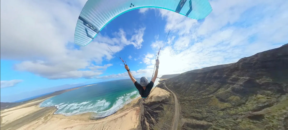 Pilot under a teal wing soaring a coastal ridge above a long sand beach and breaking surf