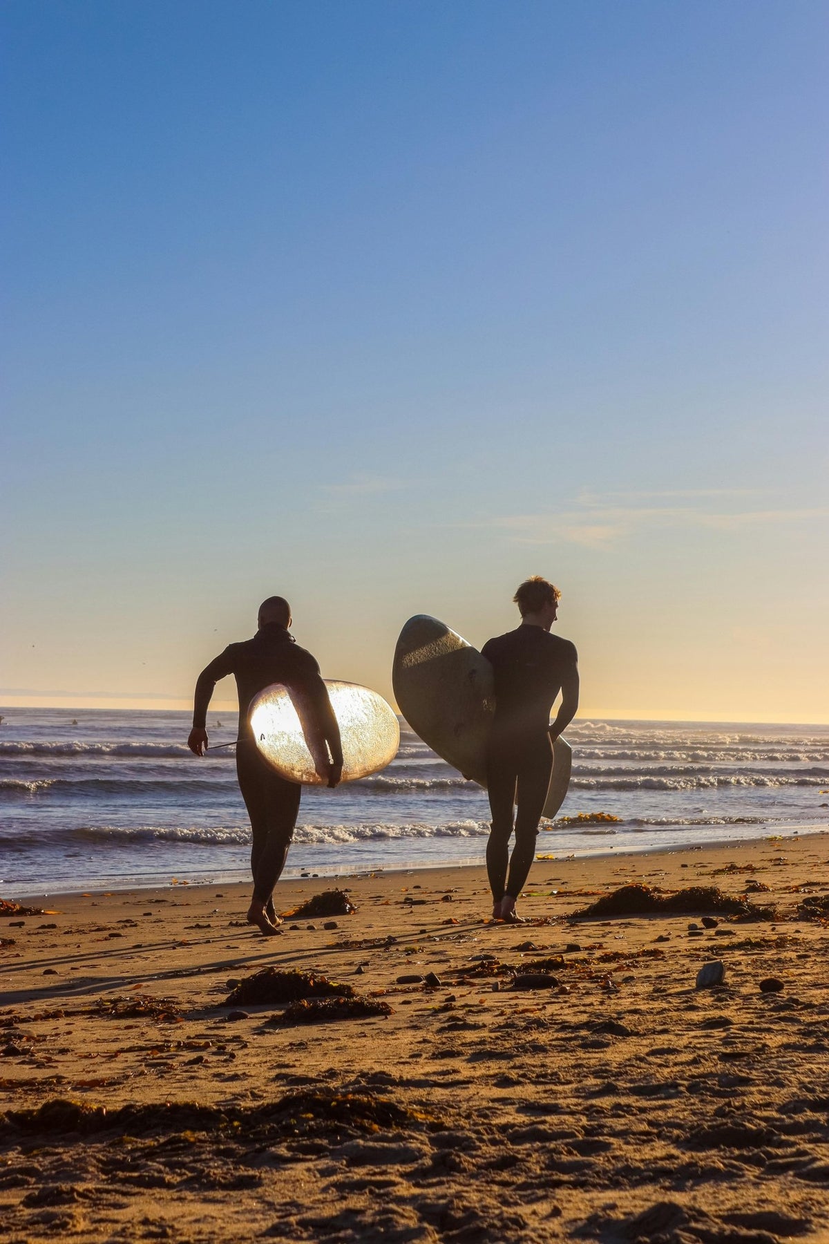 Two surfers carrying boards toward the Pacific at dawn, silhouettes against morning light