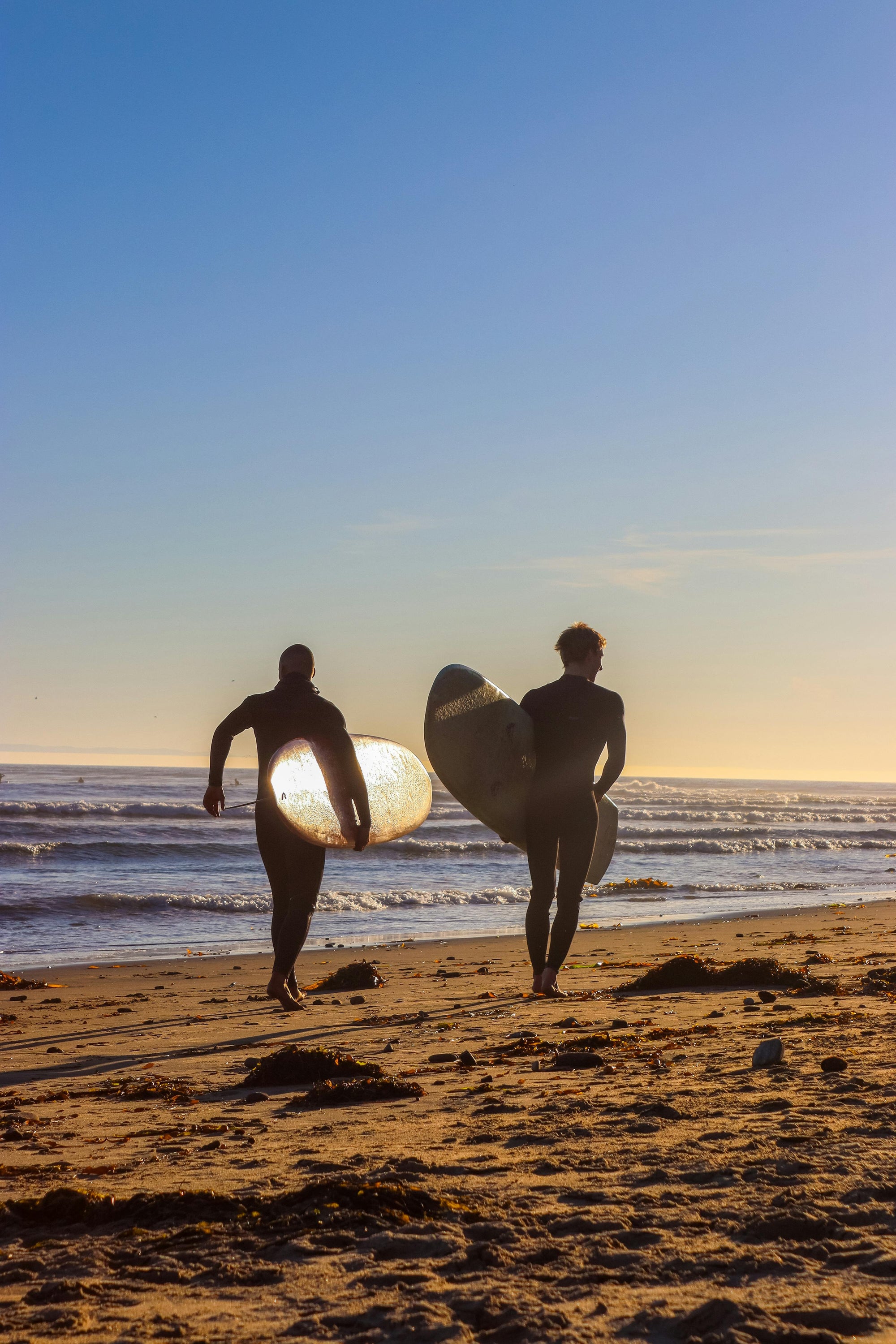 Two surfers carrying boards toward the Pacific at dawn, silhouettes against morning light